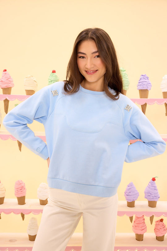 A woman in the Ivy City Co Casey Crewneck Sweatshirt in Light Blue and white pants smiles in front of a display of colorful, decorative ice cream cones on pastel shelves.