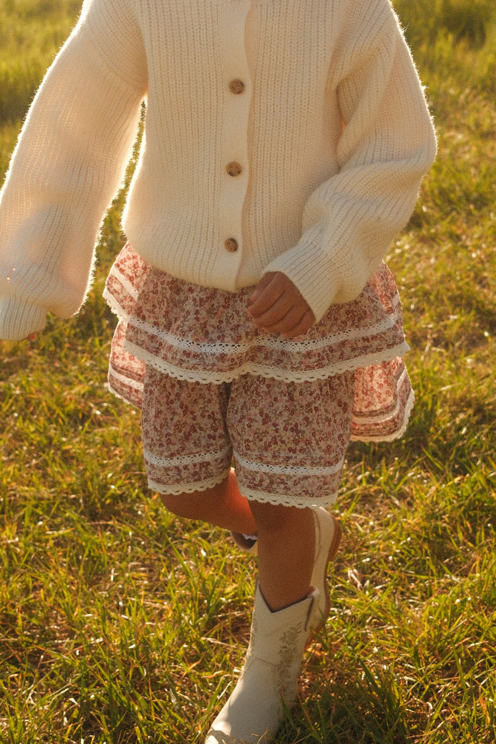 A child wearing a white sweater, white boots, and the Ivy City Co Mini Annalise Skirt in Philocaly Floral walks on grass in warm sunlight.