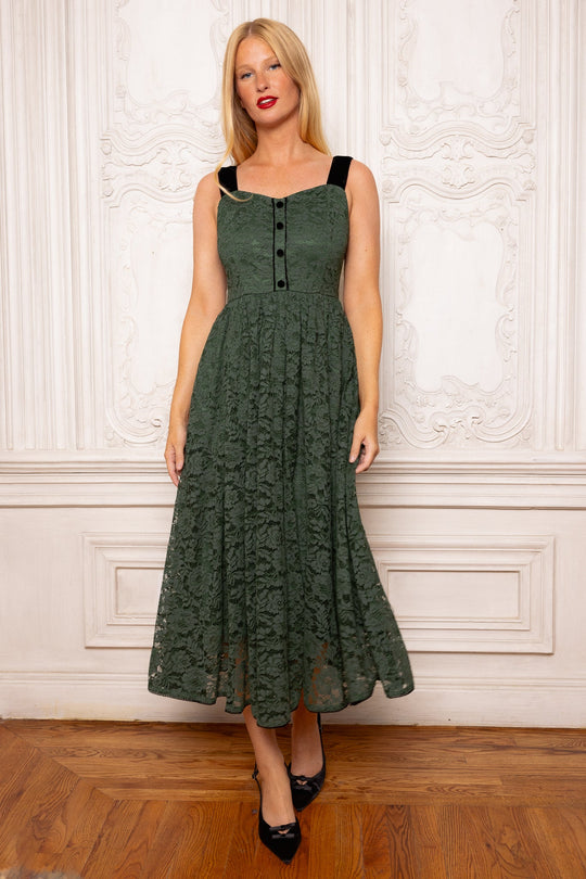 A woman wears the Ivy City Co Penny Tank Midi Dress in green lace—a sleeveless, forest-green midi dress with adjustable black straps—as she stands on a wooden floor before an ornate white wall, paired with black heels and red lipstick.