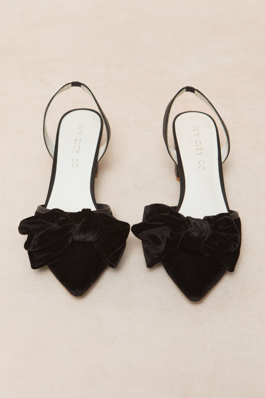 The Eloise Bow Heel in Black by Ivy City Co, featuring pointed toes and large velvet bows on the front, is elegantly displayed on a light beige background—these sophisticated heels effortlessly blend style and grace.