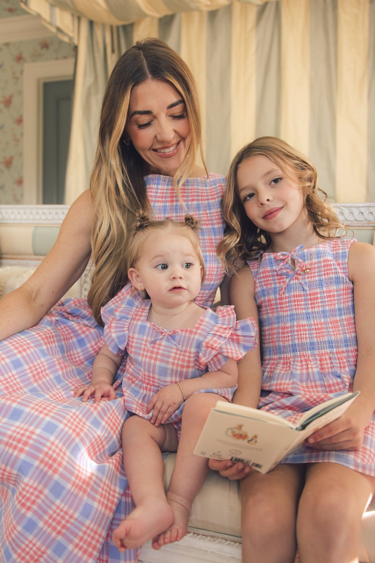A woman and two young girls sit on a couch in a cozy, well-lit room, looking happy and relaxed. One girl wears the Ivy City Co Baby Ana Romper in Blossom Plaid and holds an open book.