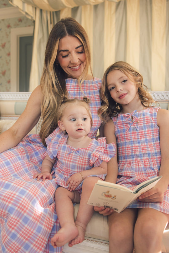 A woman and two young girls sit on a couch in a cozy, well-lit room, looking happy and relaxed. One girl wears the Ivy City Co Baby Ana Romper in Blossom Plaid and holds an open book.