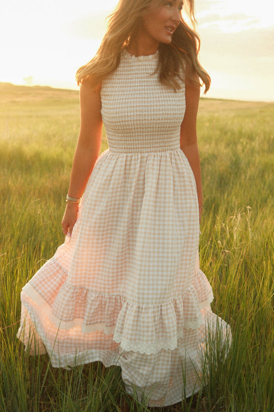A woman in the Presley Sleeveless Dress in Cream Gingham by Ivy City Co stands in a sunlit field of tall grass, soft sunlight highlighting her flowing hair and the ruffled skirt of her maternity-friendly dress.