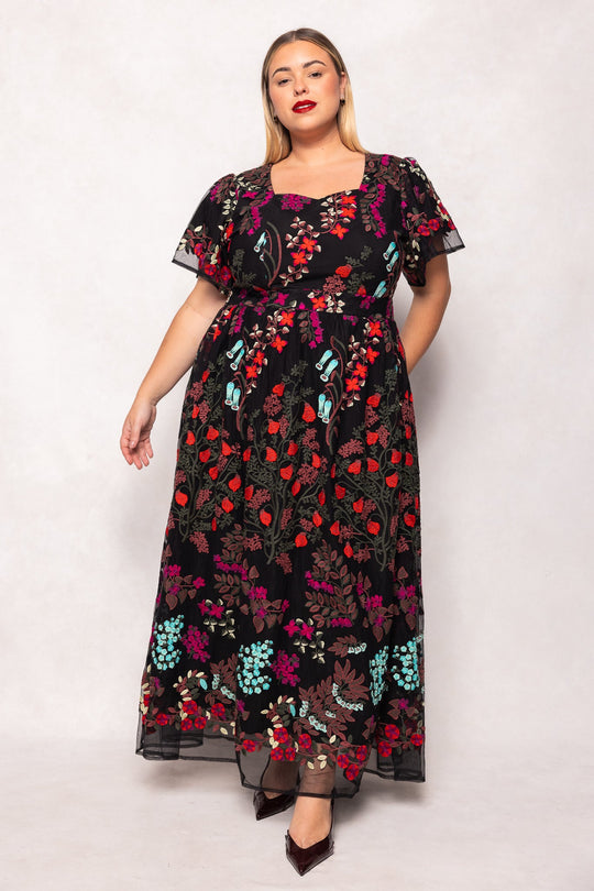 A woman poses in the Ivy City Co Flora Dress in Moonlit Blossom, a flowing maxi dress adorned with embroidered florals, colorful stitching, short sleeves, and a square neckline. She wears red lipstick and dark shoes while standing against a plain light background. This exquisite piece beautifully blends classic charm with vibrant modern accents.