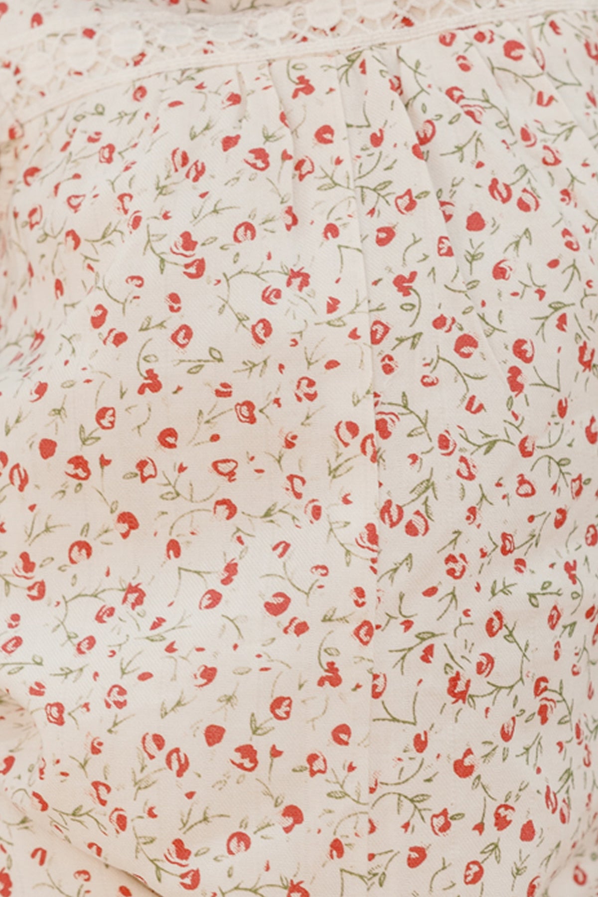 Close-up of the Ivy City Co Baby Mirabelle Romper in Cream Ditsy Floral, showcasing its cream-colored cotton fabric adorned with a delicate pattern of small red flowers and green stems, highlighted by gentle gathering and a lace trim along the top edge.