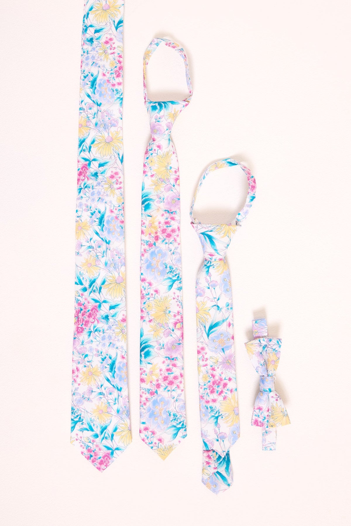 Four neckwear pieces from Ivy City Co’s Ties in Pretty Plumeria collection are arranged vertically on a light background: two ties—including an adorable Easter-themed design—one child’s tie, and one bow tie, all adorned with pastel pink, blue, and yellow plumeria flowers. Perfect for spring celebrations, these accessories add a cheerful touch to any occasion.