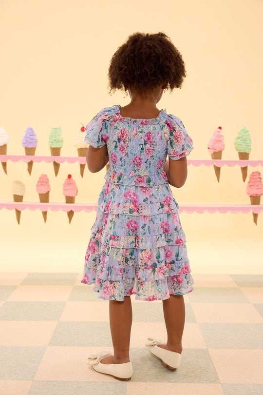 A young girl wearing the Ivy City Co Mini Winnie Dress in Peony Blossoms, featuring a smocked bodice, stands on a checkered floor in front of a pastel yellow wall decorated with ice cream cone cutouts.