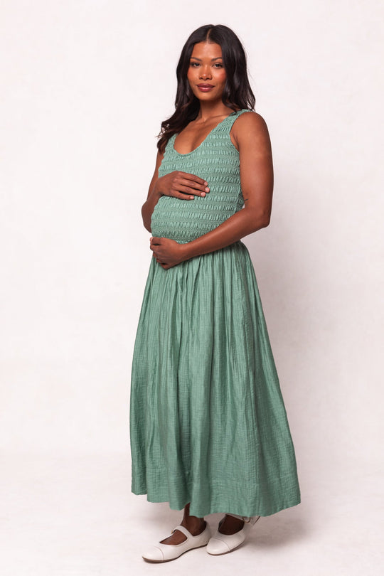 A woman with long dark hair stands against a white background, gently smiling and resting her hands on her belly as she models the Marlow Dress in Green by Ivy City Co—a light green midi dress featuring a smocked bodice and a maternity nursing-friendly design. She completes the look with white shoes.