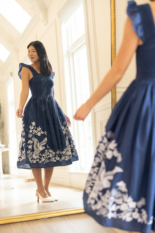 Wearing the Finch Dress in Blue by Ivy City Co, a woman with white embroidered details admires her reflection in a large mirror. She stands on light wooden flooring in a bright room with white walls and windows.