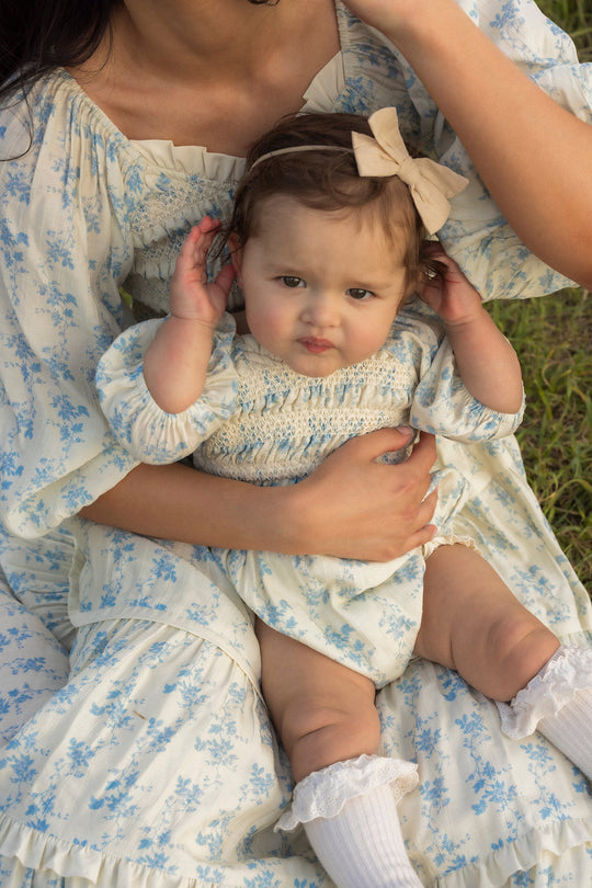 A baby wearing the Ivy City Co Baby Roselyn Romper in Ivory Porcelain Fleur, featuring a cream fabric with blue floral print and a beige bow, sits on a woman’s lap in matching outfits outdoors on the grass.