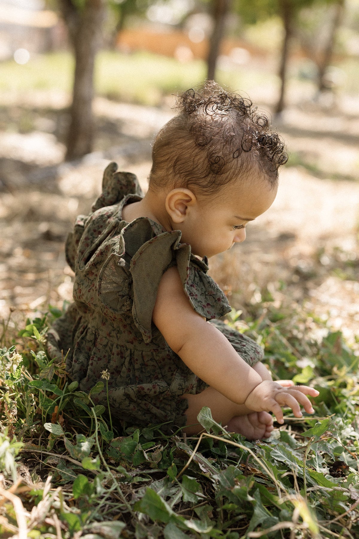 A baby with curly hair sits on the grass outdoors, wearing the Ivy City Co Baby Hattie Romper in Woodland Floral with flutter sleeves. The baby looks down and gently touches the leaves, surrounded by sunlight and trees in the background.
