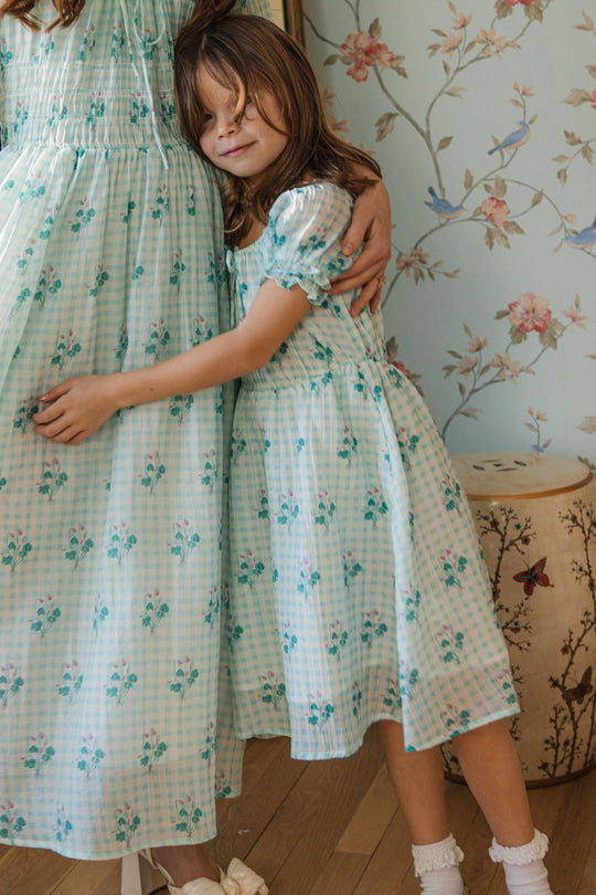A young girl wearing the Mini Jessie Dress in Bloom Gingham by Ivy City Co hugs an adult dressed in a matching outfit. They stand on a wooden floor beside a floral-patterned wall, bathed in soft light, with a decorative stool nearby.