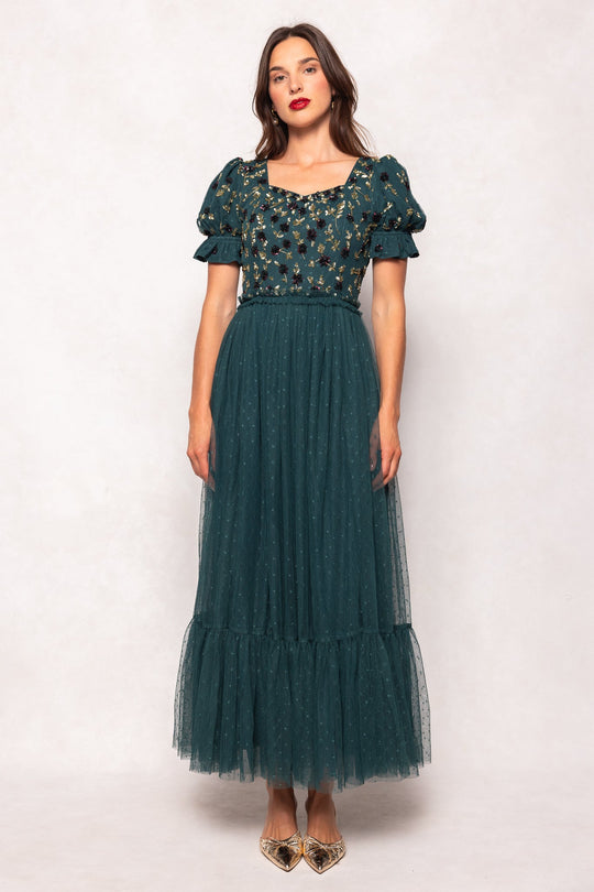A woman wearing the Ivy City Co Adara Dress in Green—an ankle-length gown featuring a tulle skirt, puff sleeves, and floral embroidery on the bodice—paired with gold pointed shoes, stands against a light background.