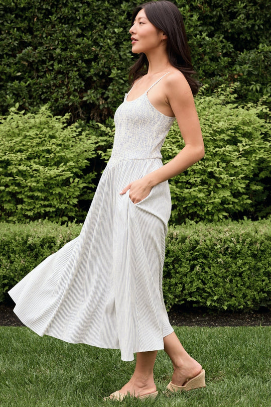 A woman wearing the Ivy City Co Maxwell Dress in Blue & White Pinstripe—an airy, sleeveless, ankle-length maternity dress—walks on grass with greenery in the background, gazing ahead with one hand in her pocket.