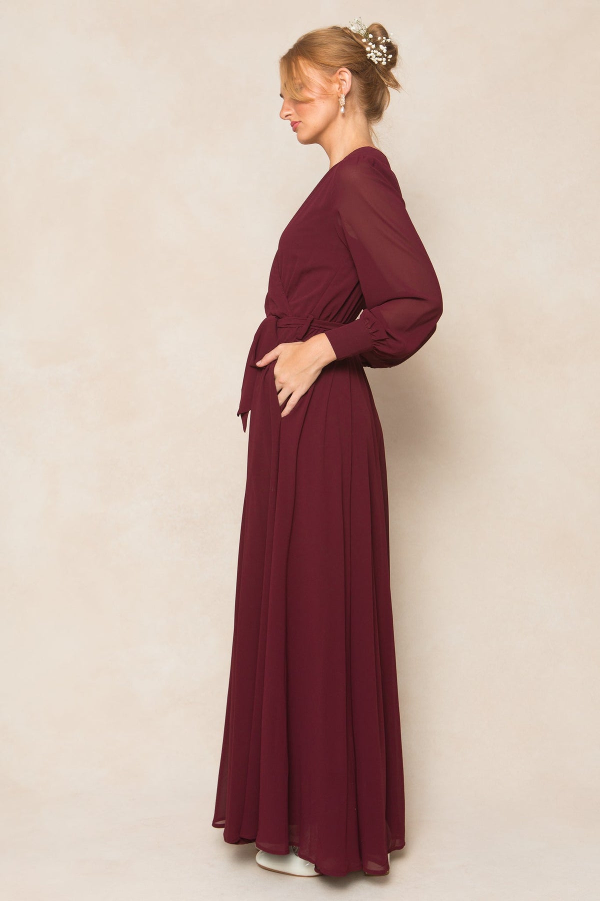 A woman stands in profile against a light beige background, her hair styled in an elegant updo and one hand resting on her waist, wearing the Ivy City Co Andie Dress in Wine Chiffon—a long-sleeved, floor-length bridesmaid gown.