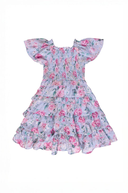The Ivy City Co Mini Winnie Dress in Peony Blossoms features short sleeves, ruffled details, a smocked bodice, and a tiered skirt, all adorned with a pink and green peony floral pattern on a light purple background.