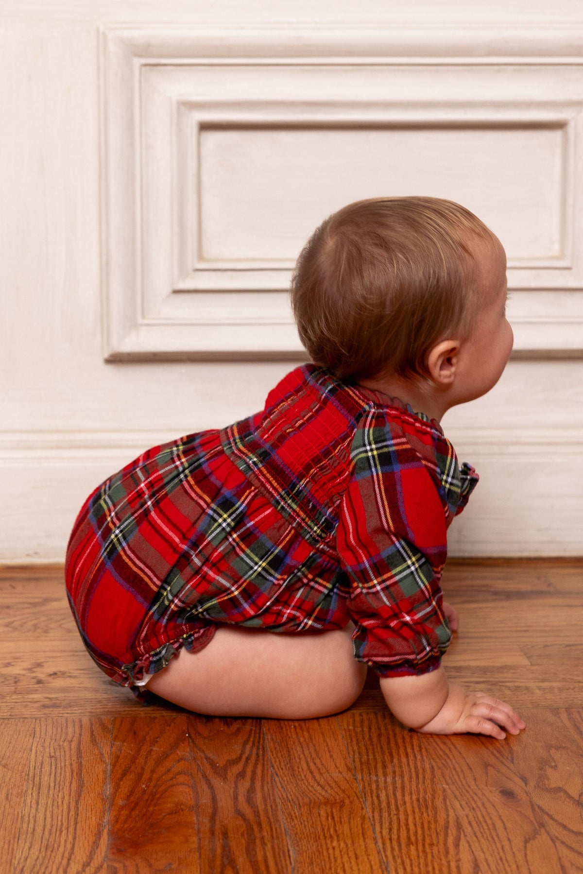 Dressed in the Ivy City Co Baby Madeline Romper in Holiday Plaid, a baby sits on a wooden floor facing away from the camera and gazing toward a white paneled door.
