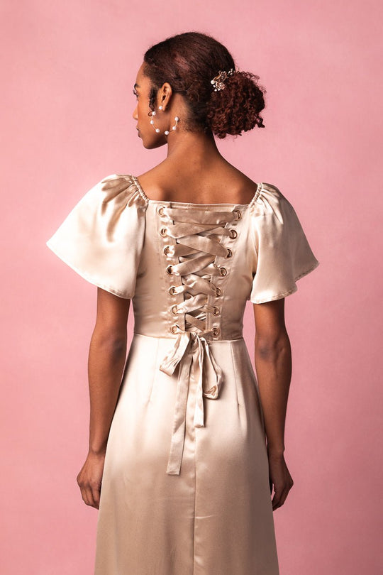 A woman with curly hair styled in an updo, adorned with pearl hairpins, stands against a pink background facing away from the camera. She wears the Ivy City Co Maddie Dress in Champagne Satin, featuring short sleeves and a lace-up back.