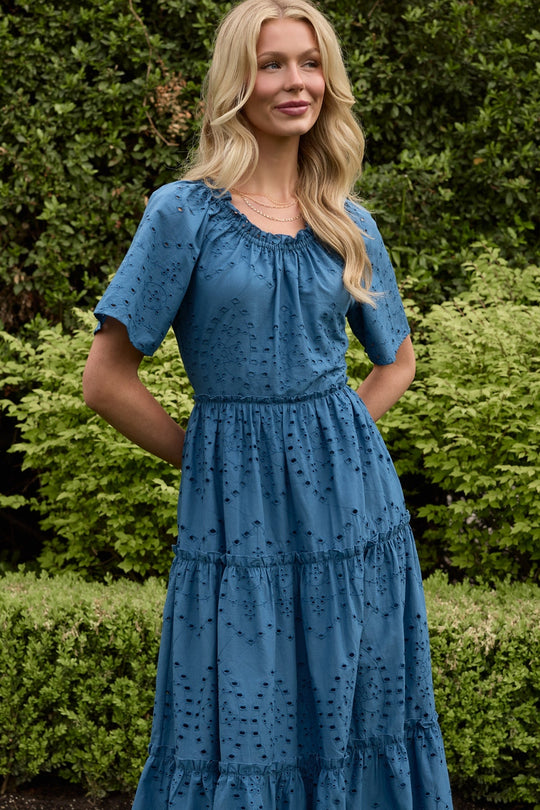 A woman with long blonde hair stands outdoors in front of green bushes, wearing the Ivy City Co Etta Dress in Cobalt—a vibrant blue midi dress featuring short sleeves, eyelet details, a tiered silhouette, and convenient pockets. She smiles as she casually tucks her hands into her dress pockets, embodying the effortless charm of this summer staple.