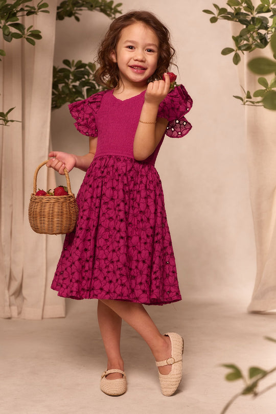 A smiling young girl wearing the Ivy City Co Mini Hattie Dress in Magenta holds a small basket of strawberries in one hand and a strawberry in the other, surrounded by lush green plants.