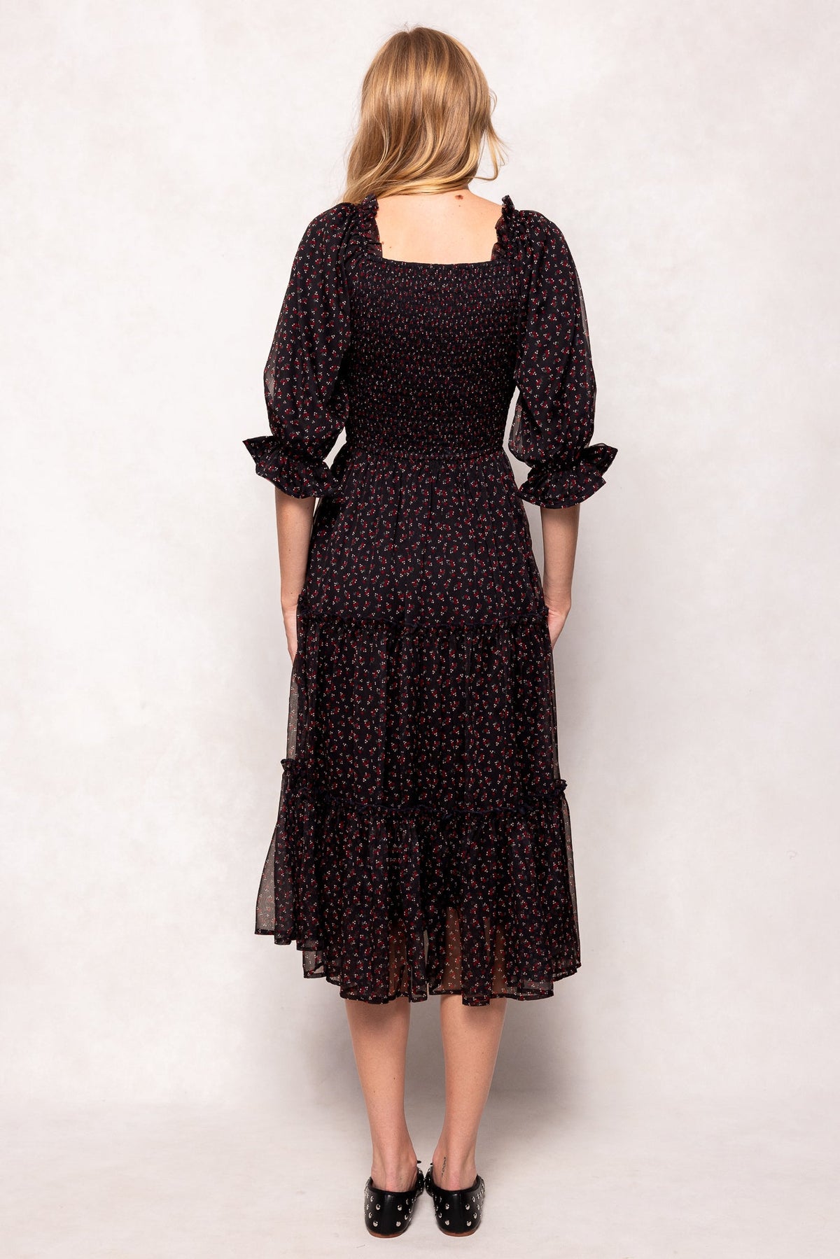 A woman stands facing away, wearing the Ivy City Co Madeline Dress in Black Ditsy Floral—a nursing-friendly and maternity-friendly midi dress featuring long sleeves and a smocked bodice—paired with black shoes, against a light background.