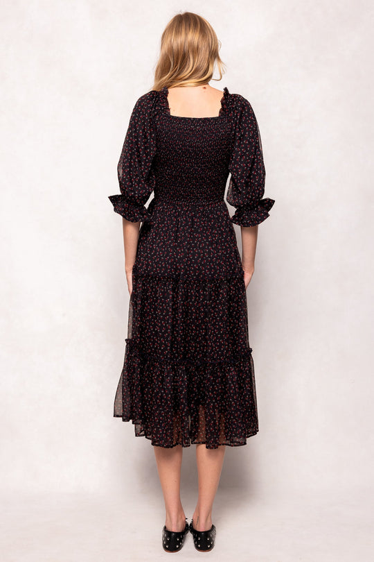 A woman stands facing away, wearing the Ivy City Co Madeline Dress in Black Ditsy Floral—a nursing-friendly and maternity-friendly midi dress featuring long sleeves and a smocked bodice—paired with black shoes, against a light background.