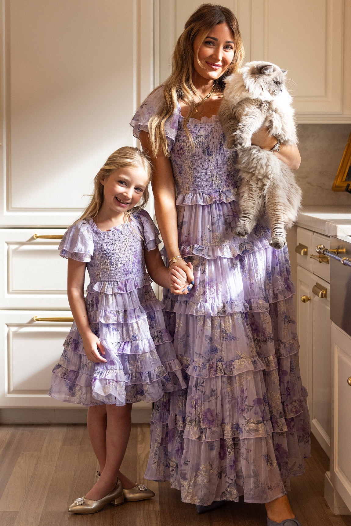 A woman and a girl, both wearing matching Ivy City Co Mini Winnie Dresses in Lavender Aquarelle Fleur with smocked bodices and ruffle tiered skirts, stand in a kitchen holding hands. The woman holds a fluffy white cat as sunlight streams in, creating a warm and cozy atmosphere.