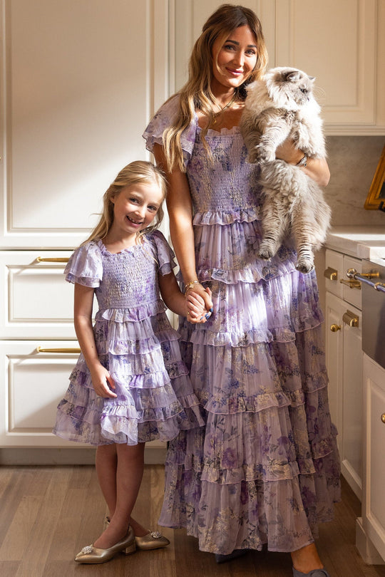 A woman and a girl, both wearing matching Ivy City Co Mini Winnie Dresses in Lavender Aquarelle Fleur with smocked bodices and ruffle tiered skirts, stand in a kitchen holding hands. The woman holds a fluffy white cat as sunlight streams in, creating a warm and cozy atmosphere.
