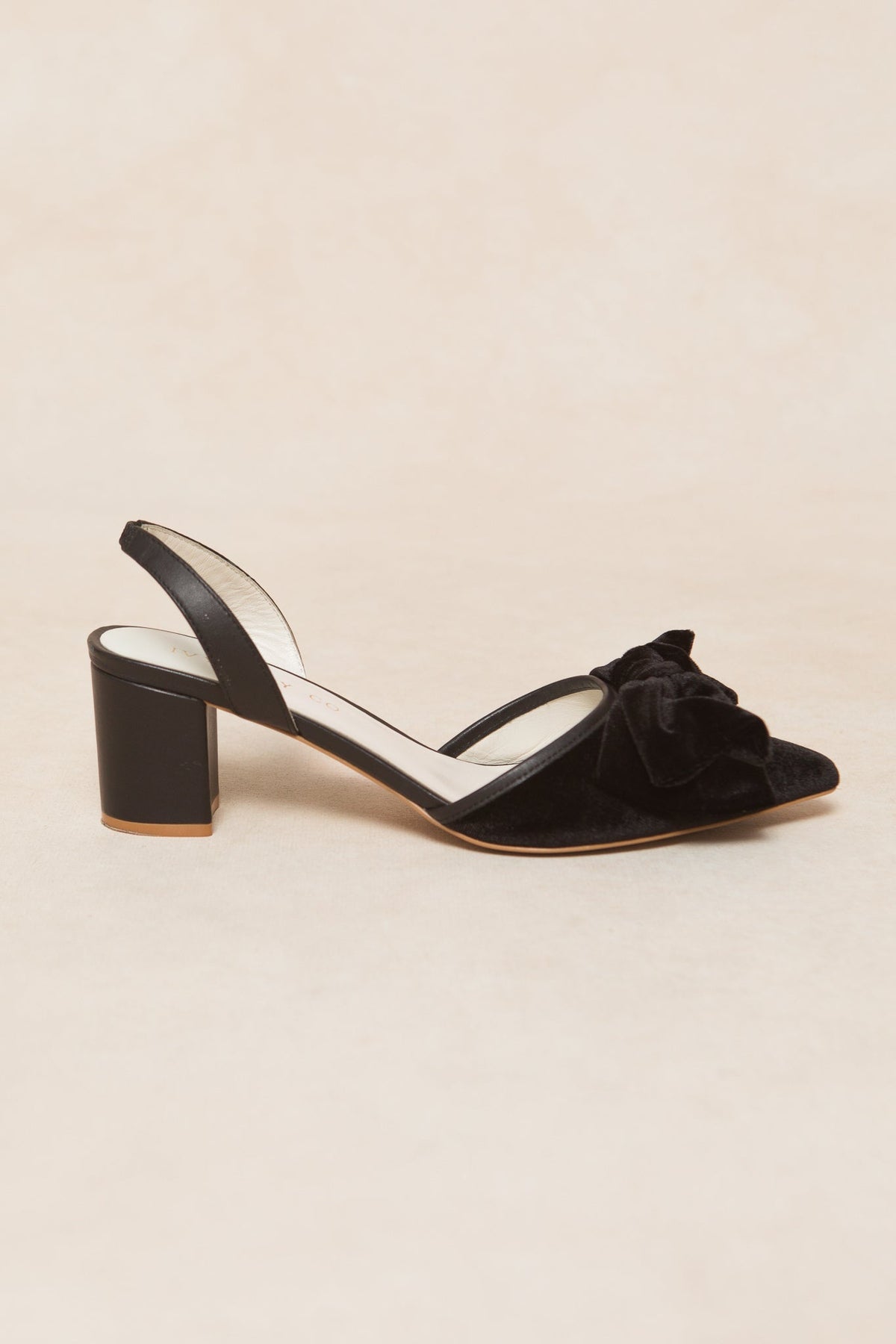 The Eloise Bow Heel in Black by Ivy City Co is a pair of slingback heels crafted from suede, showcasing a pointed toe, sturdy block heel, and a decorative bow on the front, elegantly displayed on a light beige background.