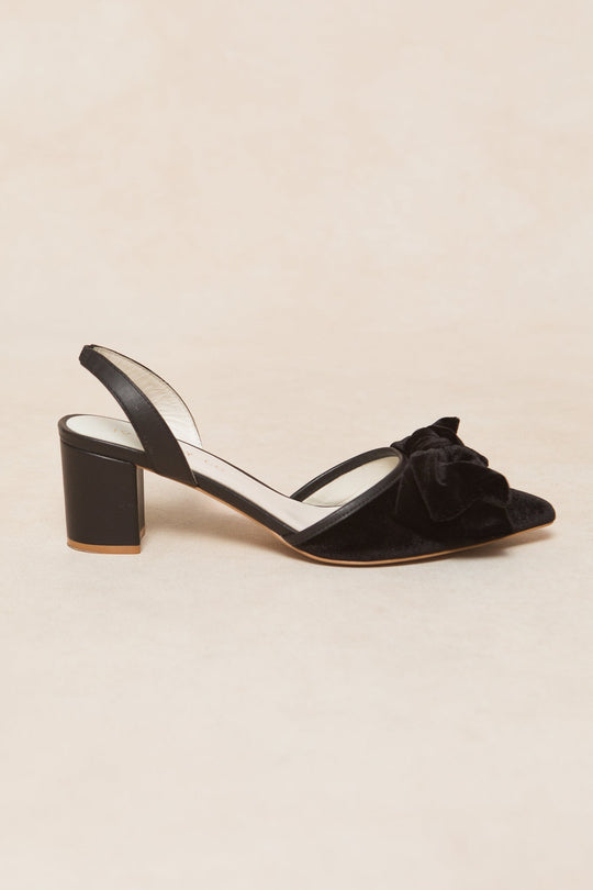 The Eloise Bow Heel in Black by Ivy City Co is a pair of slingback heels crafted from suede, showcasing a pointed toe, sturdy block heel, and a decorative bow on the front, elegantly displayed on a light beige background.