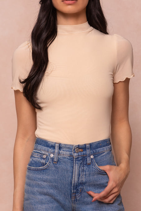 A woman wears the Ivy City Co Ava Top in Cream—a short-sleeve, maternity-friendly mock neckline top—with high-waisted blue jeans. She stands with one hand in her pocket against a light pink background, her long dark hair worn loose.
