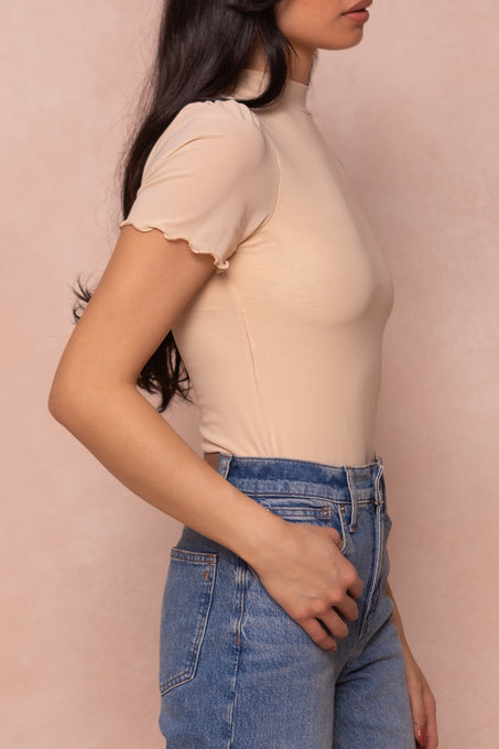 A woman with long dark hair wears the Ava Top in Cream by Ivy City Co and high-waisted blue jeans, standing against a soft pink background, shown from shoulders to hips in a side profile.