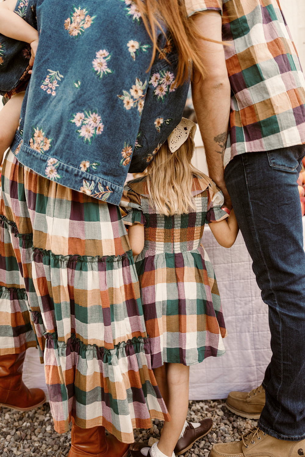 A family stands close together outdoors, dressed in colorful plaid outfits and boots. One young girl wears the Ivy City Co Mini Alice Dress in Maize Gingham, featuring puff sleeves and a smocked bodice. A hair bow adorns her head as she holds an adult’s hand, their faces not visible.