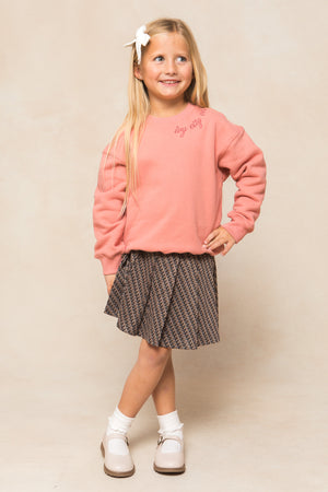 A young girl with long blonde hair and a white bow smiles, wearing the Ivy City Co Mini Ivy Sweatshirt in Pink with a patterned skirt, white socks, and beige shoes, standing with one hand on her hip against a light neutral background.