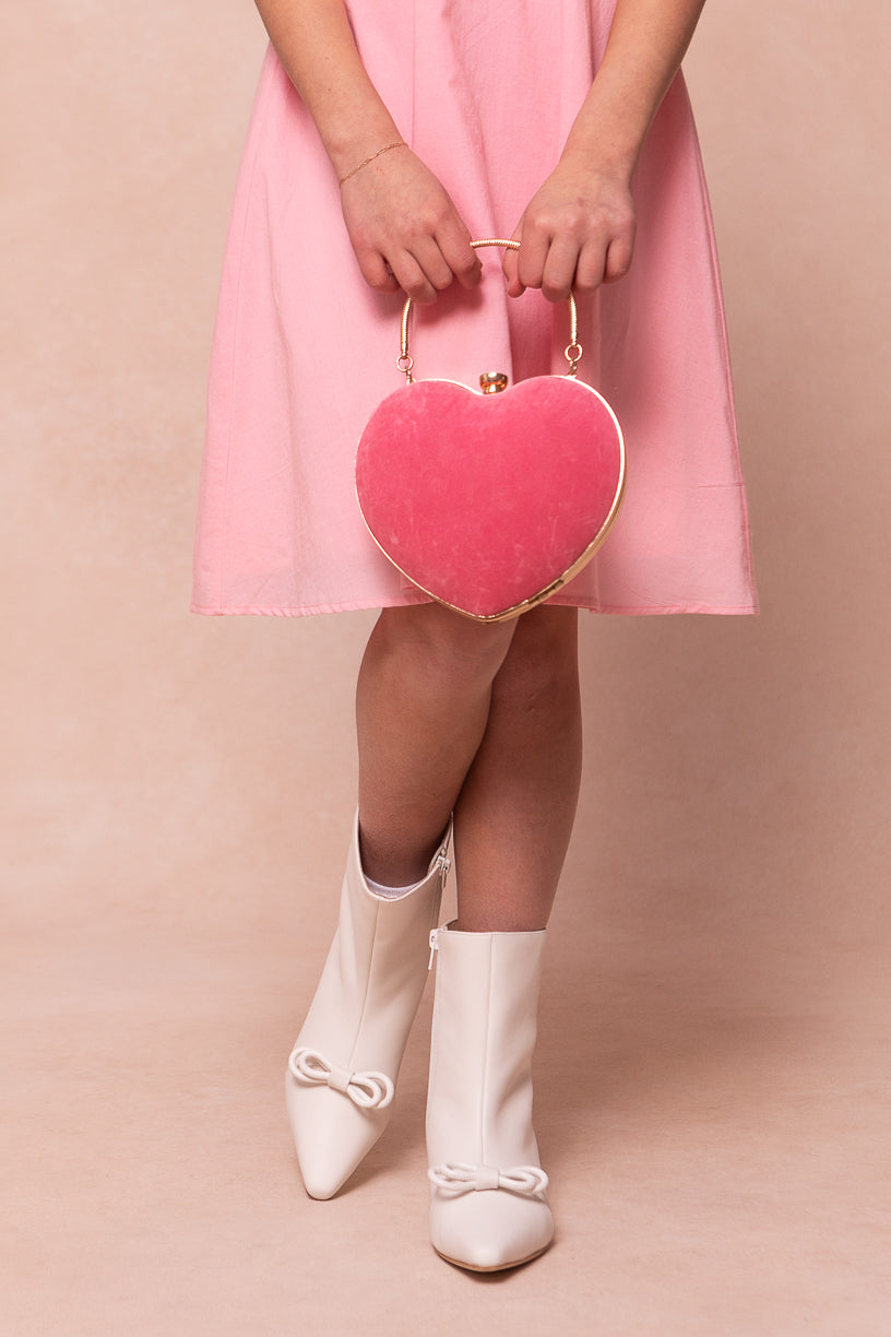 A person in a pink dress stands with legs crossed, holding a pink heart-shaped handbag and wearing Ivy City Cos Mini Ivy Bow Ankle Boot in White. The background is soft beige.