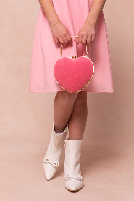 A person in a pink dress stands with legs crossed, holding a pink heart-shaped handbag and wearing Ivy City Cos Mini Ivy Bow Ankle Boot in White. The background is soft beige.