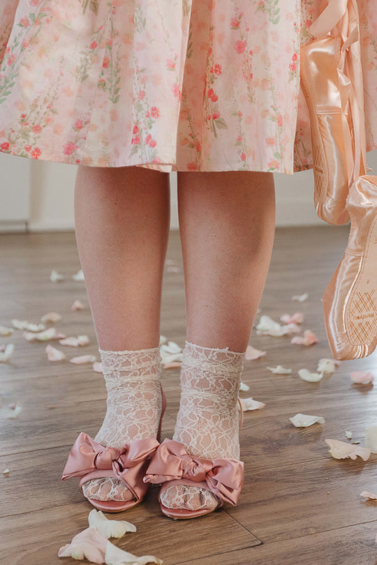 A person in a pink floral dress stands on a wooden floor scattered with petals, wearing lace socks and the Ivy City Co Ivy Bow Heel in Pink Satin; ballet pointe shoes hang nearby.