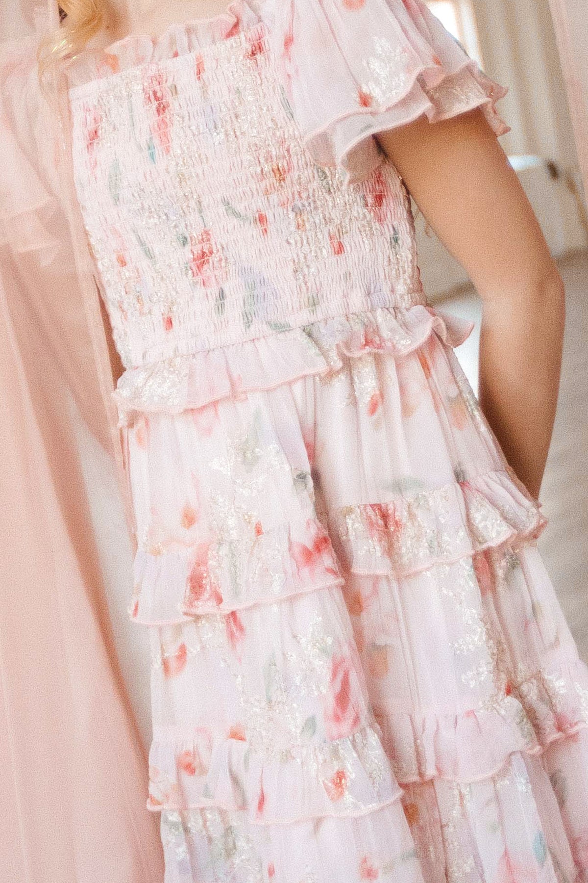 A close-up of a person wearing the Mini Winnie Dress in Pink Floral by Ivy City Co, featuring a smocked bodice, tiered ruffles, glittery accents, and airy fabric for a soft, romantic look.