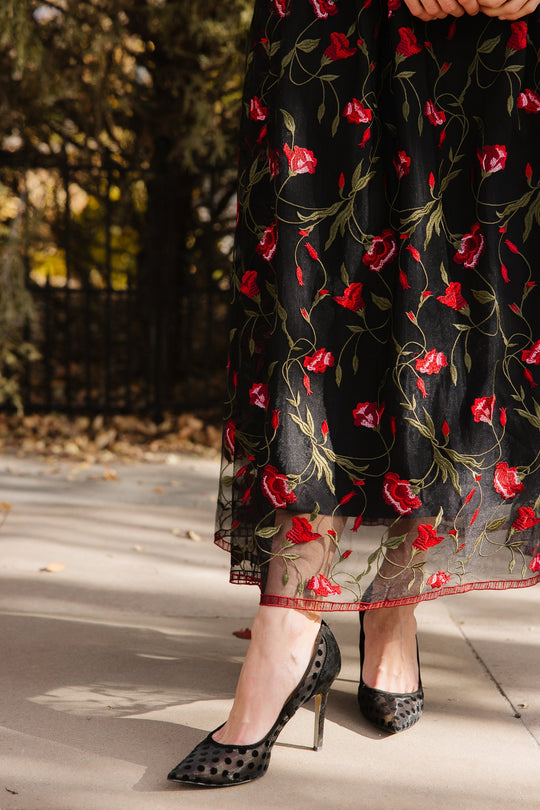 A person in the Ivy City Co Flora Dress in Romantic Roses—a black dress featuring a black mesh overlay and embroidered red roses—stands outside, wearing black high heels with a mesh polka dot pattern.