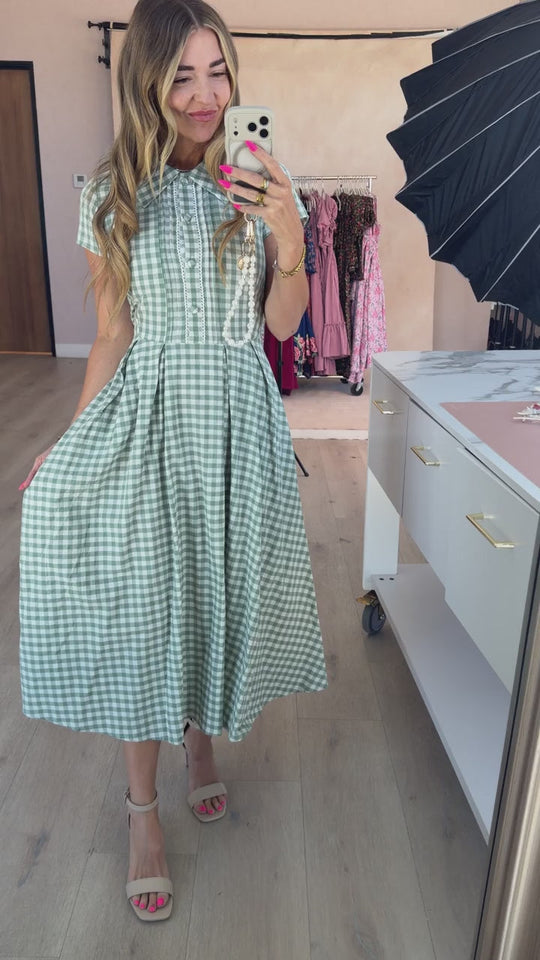 A woman takes a mirror selfie wearing the Amber Dress in Sage Gingham by Ivy City Co, featuring a pleated skirt, paired with beige sandals. She stands in a bright room with clothing racks, a white cabinet, and photography equipment visible in the background.