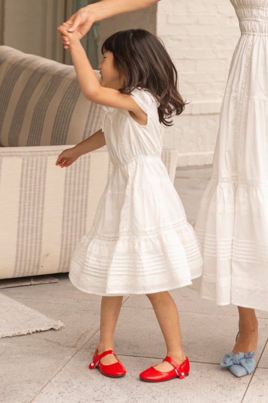 A young girl wears the Ivy City Co Mini Palermo Dress in White as she holds hands and dances indoors with an adult dressed in a coordinating summer dress.