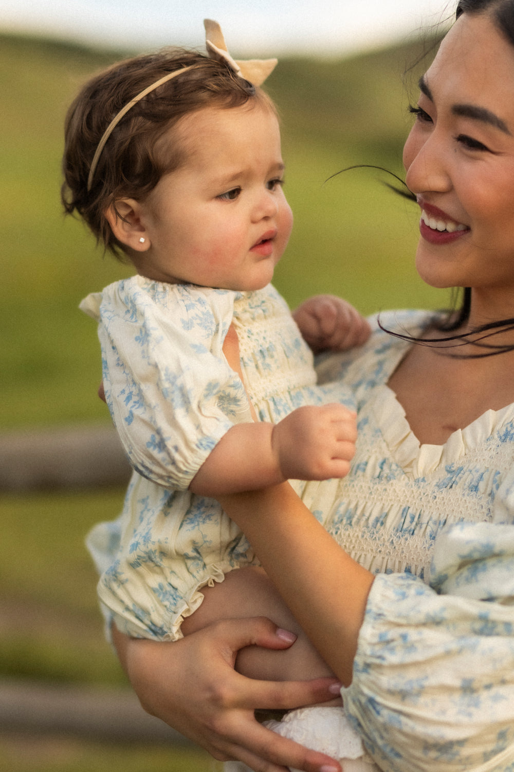 A woman smiles while holding a baby dressed in the adorable Baby Roselyn Romper in Ivory Porcelain Fleur from Ivy City Co, complete with a matching floral headband. They are outdoors with blurred greenery in the background.