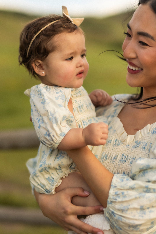 A woman smiles while holding a baby dressed in the adorable Baby Roselyn Romper in Ivory Porcelain Fleur from Ivy City Co, complete with a matching floral headband. They are outdoors with blurred greenery in the background.
