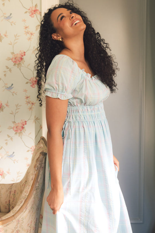 A woman with curly hair, wearing the Thalia Dress in Pastel Plaid by Ivy City Co, stands smiling and looking up, bathed in soft sunlight beside a floral wallpapered wall and a patterned chair.