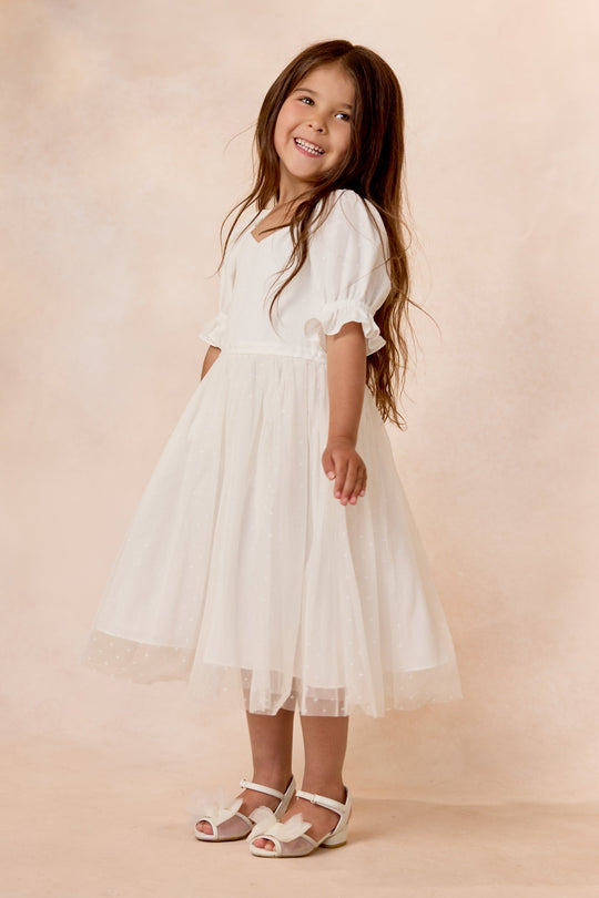 A young girl stands smiling against a soft beige background, wearing the Ivy City Co Mini Ballerina Dress in White—a knee-length dress with puffed sleeves and a delicate tulle skirt—paired with white sandals.