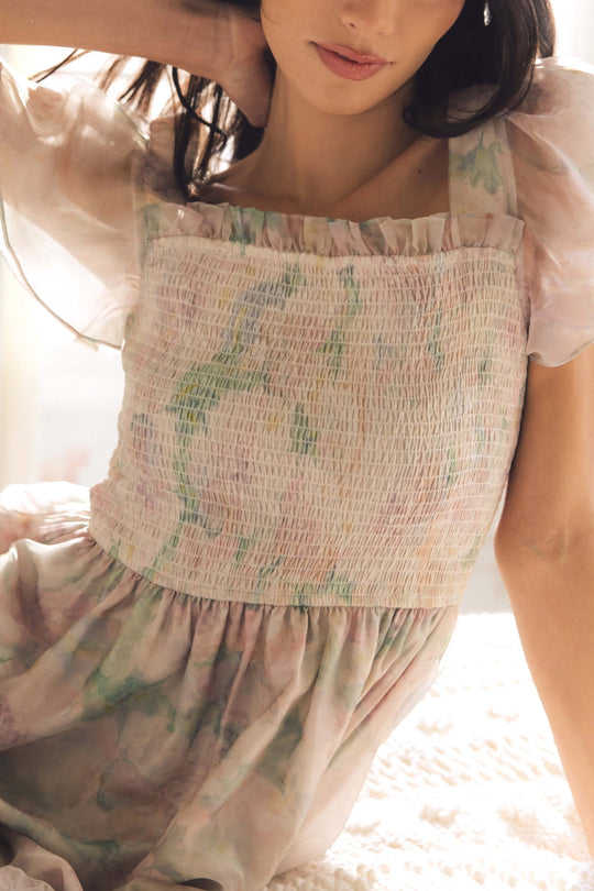 A woman wearing the Rhiannon Dress in Painted Bloom by Ivy City Co, featuring pastel watercolor florals, smocking, and puff sleeves, sits on a bed bathed in soft natural light. The photo highlights the upper half of her dress and her relaxed pose.