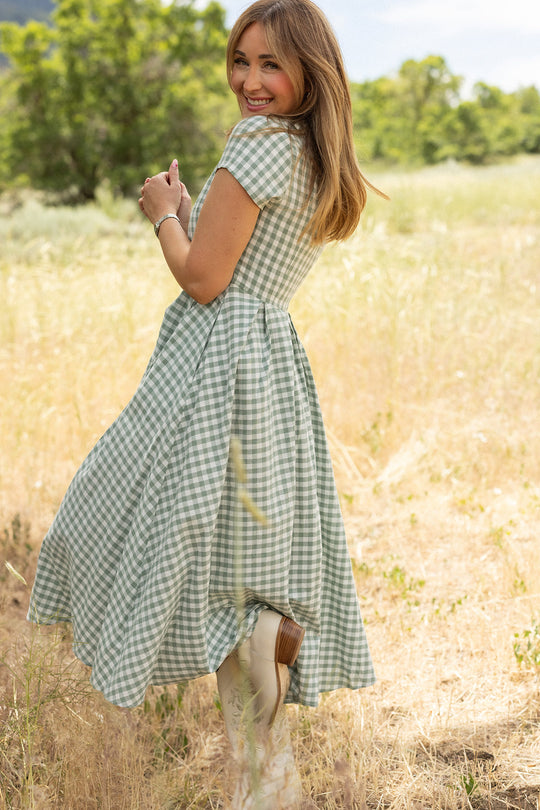 A woman stands in a sunlit field, smiling over her shoulder with her hair down, wearing the Ivy City Co Amber Dress in Sage Gingham. The cotton midi dress features a charming pleated skirt, while tall grass and trees create a beautiful backdrop.