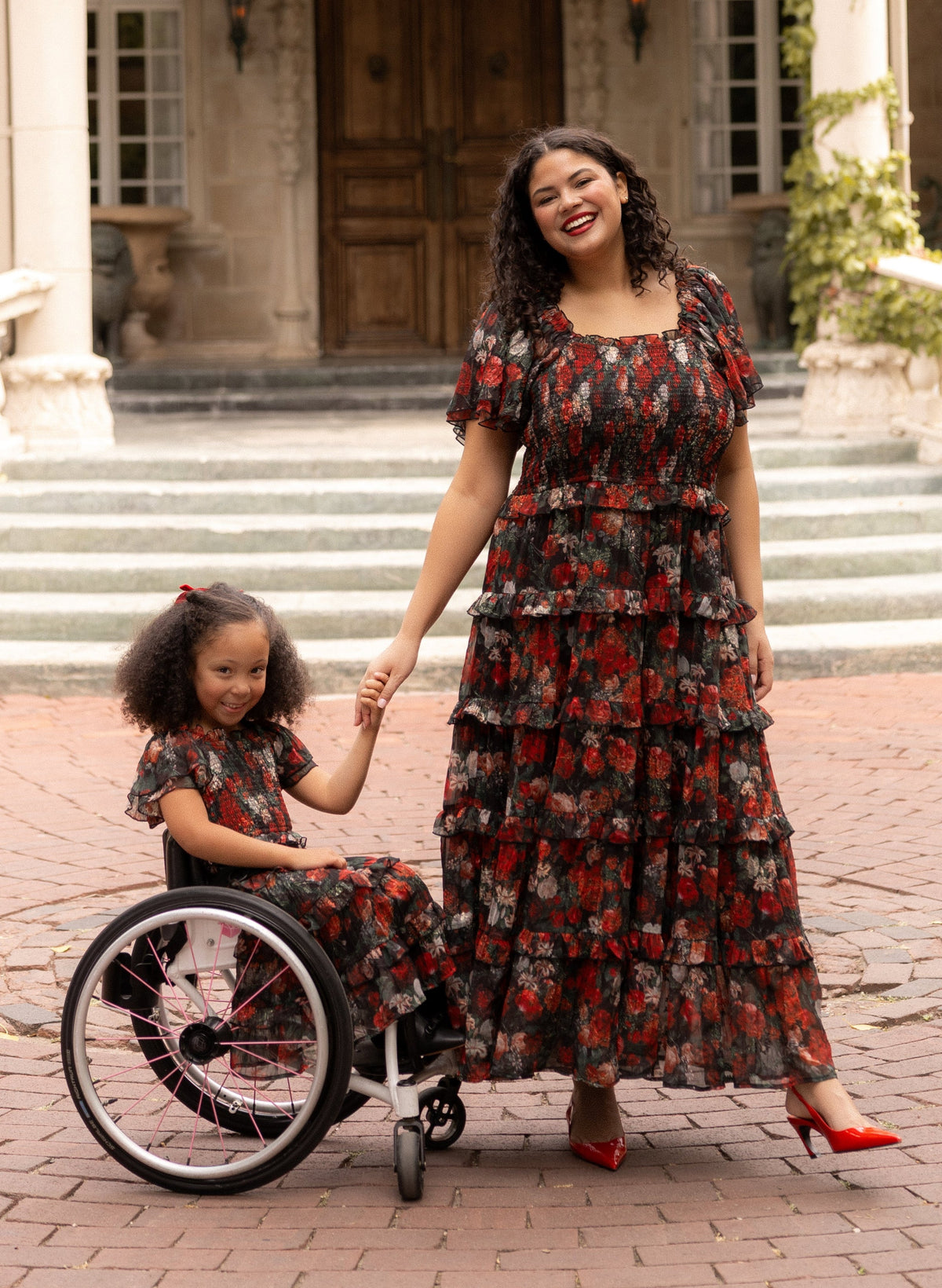 A woman and a young girl in matching floral dresses smile and hold hands. The girl, wearing the charming Mini Winnie Dress in Midnight Roses by Ivy City Co, sits in a wheelchair as they pose outdoors in front of a large building with stone steps and wooden doors—a perfect mommy-and-me dress moment.