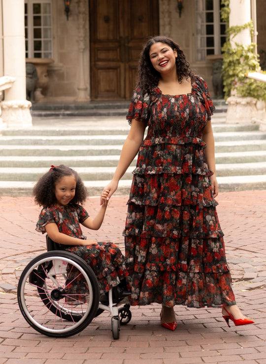 A woman and a young girl in matching floral dresses smile and hold hands. The girl, wearing the charming Mini Winnie Dress in Midnight Roses by Ivy City Co, sits in a wheelchair as they pose outdoors in front of a large building with stone steps and wooden doors—a perfect mommy-and-me dress moment.
