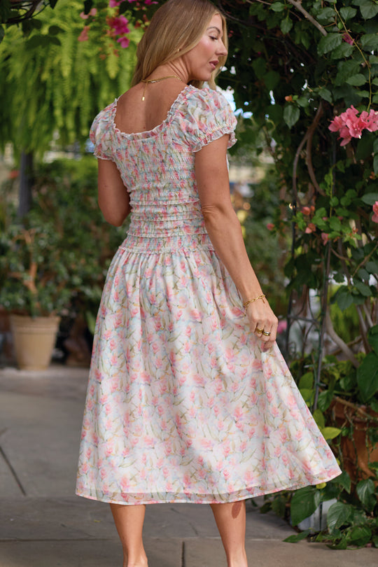 A woman wearing the Ivy City Co Kyla Dress in Watercolor Orchids stands outdoors, looking over her shoulder amid lush green plants and pink flowers.